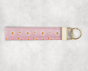 A faux leather wristlet made of a fine pink glitter fabric with a white daisy pattern. The hardware cap is a metallic gold color with a keyring on the end.